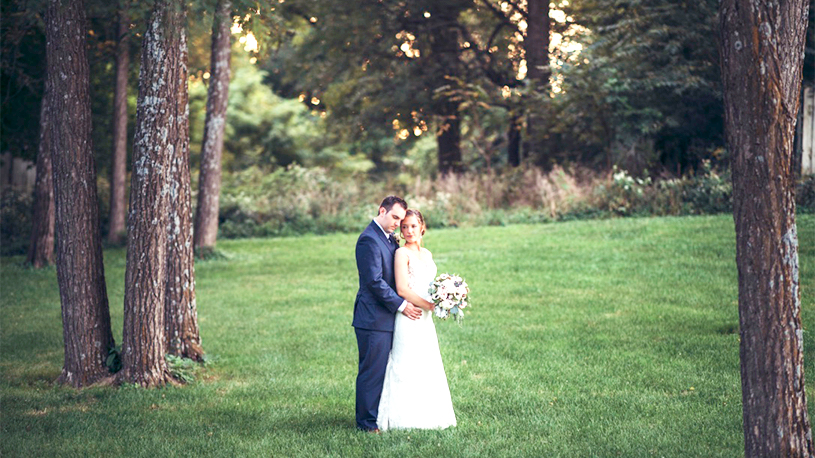 Newlywed couple posing outside on green grass between two trees.