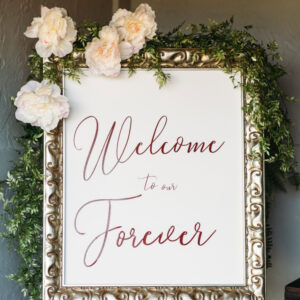"Welcome to Forever" sign framed in a gold frame with flowers and greenery draped on top.