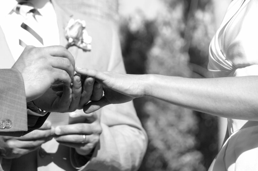 Black and white image of groom puts wedding band onto bride's finger in front of officiant.