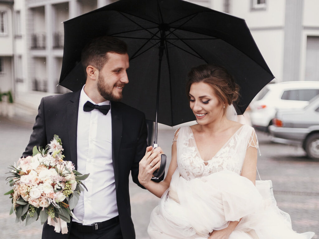 Bride holding up dress while walking in the rain with groom under an umbrella.