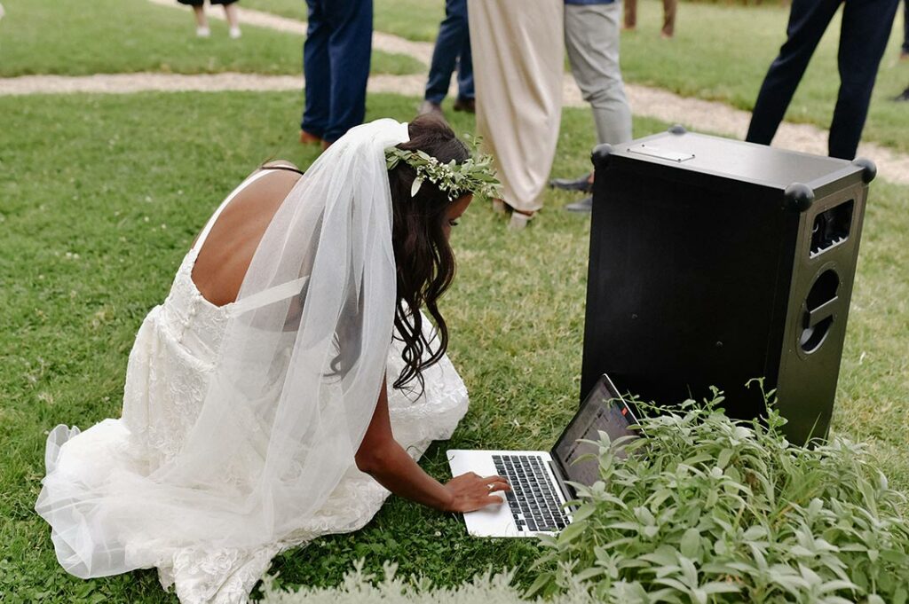 Bride bending dow to start music on a laptop for an outdoor reception.