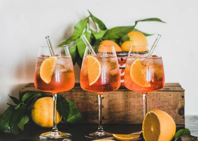 Three wine glasses filled with Spritz cocktails with a display of lemons behind them.